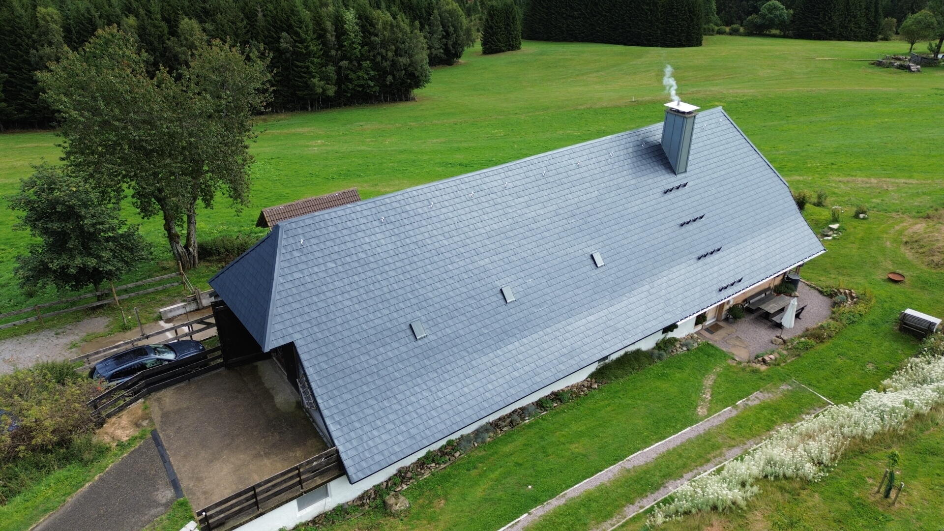 Luftaufnahme eines langen Gebäudes mit grauem Dach, aus dessen Schornstein Rauch aufsteigt, umgeben von grünem Gras, Bäumen und einer Kiesauffahrt, auf der ein Auto geparkt ist.