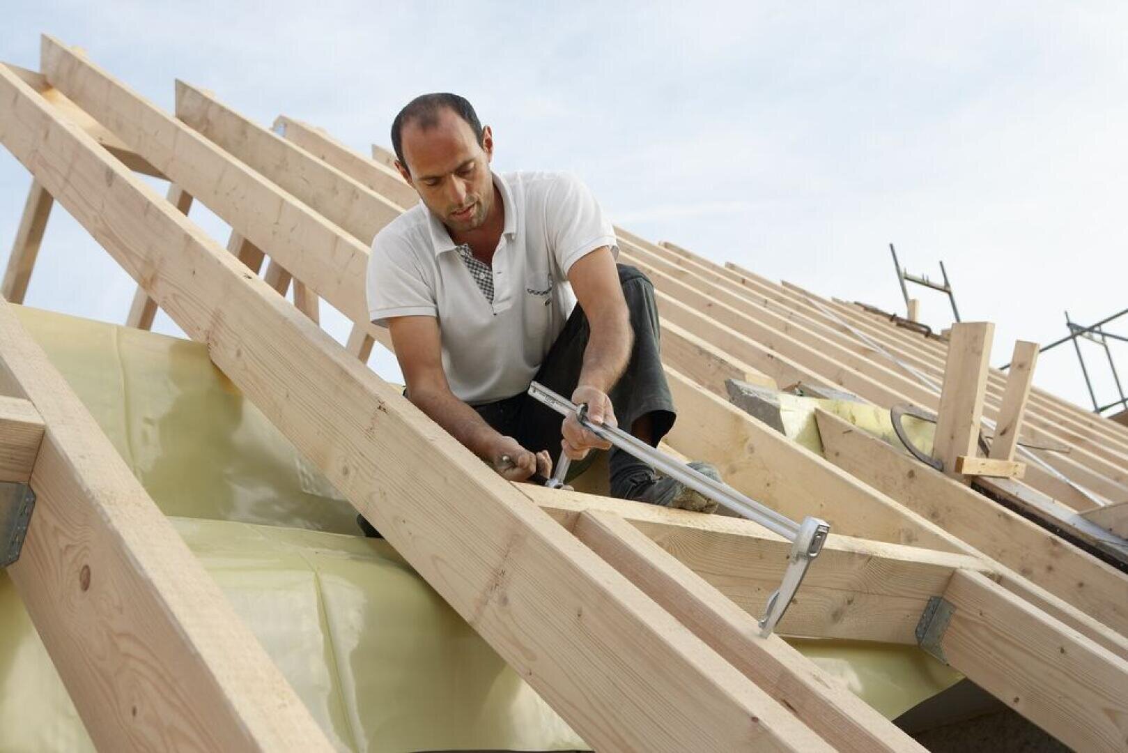 Ein Mann mit weißem Hemd kniet auf einem hölzernen Dachstuhl und befestigt die Balken mit Hammer und Nägeln, während im Hintergrund Baumaterialien und blauer Himmel zu sehen sind.