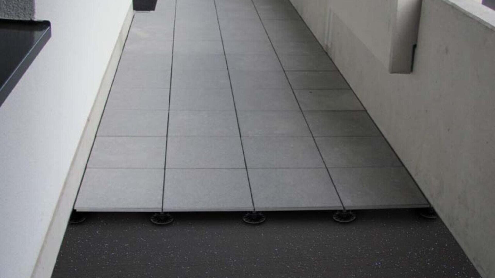 A tiled floor with several tiles lifted, revealing round supports underneath and a dark subfloor; the setting appears to be an indoor hallway or balcony with white walls.