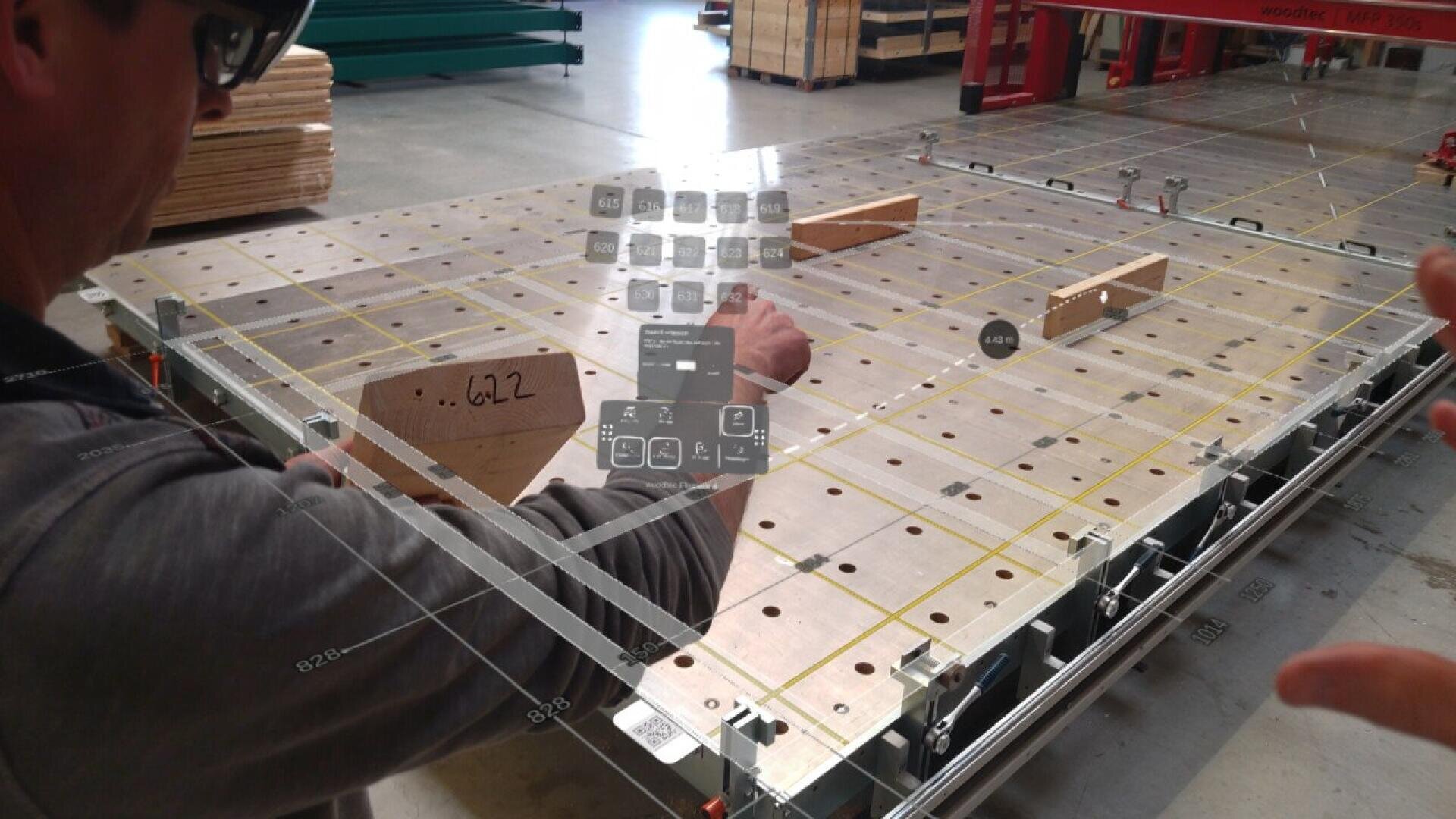 A person uses augmented reality glasses to interact with a digital interface projected over a large wooden work surface in an industrial workshop. Various icons and measurement data overlay the physical table.