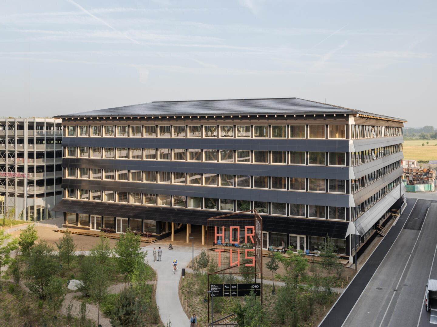 A modern, rectangular office building with large windows and a dark roof, surrounded by green space. In front of the building, the sign HORTUS is written in red letters. A road runs beside the building.