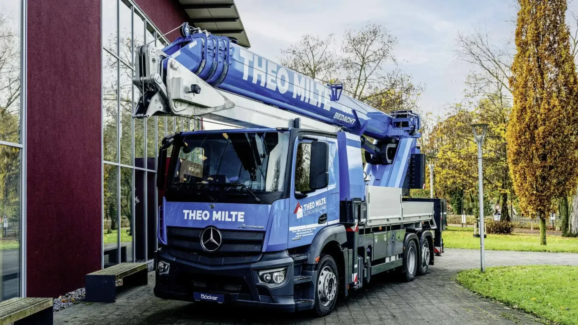 A large blue Mercedes-Benz truck with a mounted crane, labeled THEO MILTE, is parked beside a building with glass windows and surrounded by trees with autumn foliage.