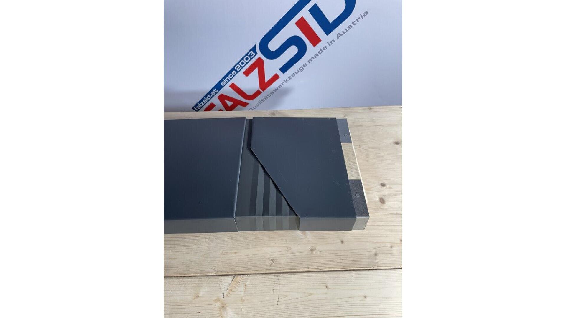 A gray rectangular metal object with a grooved section rests on a light wooden surface. In the background, part of a sign with the text “ALZSID” and “made in Austria” is visible.