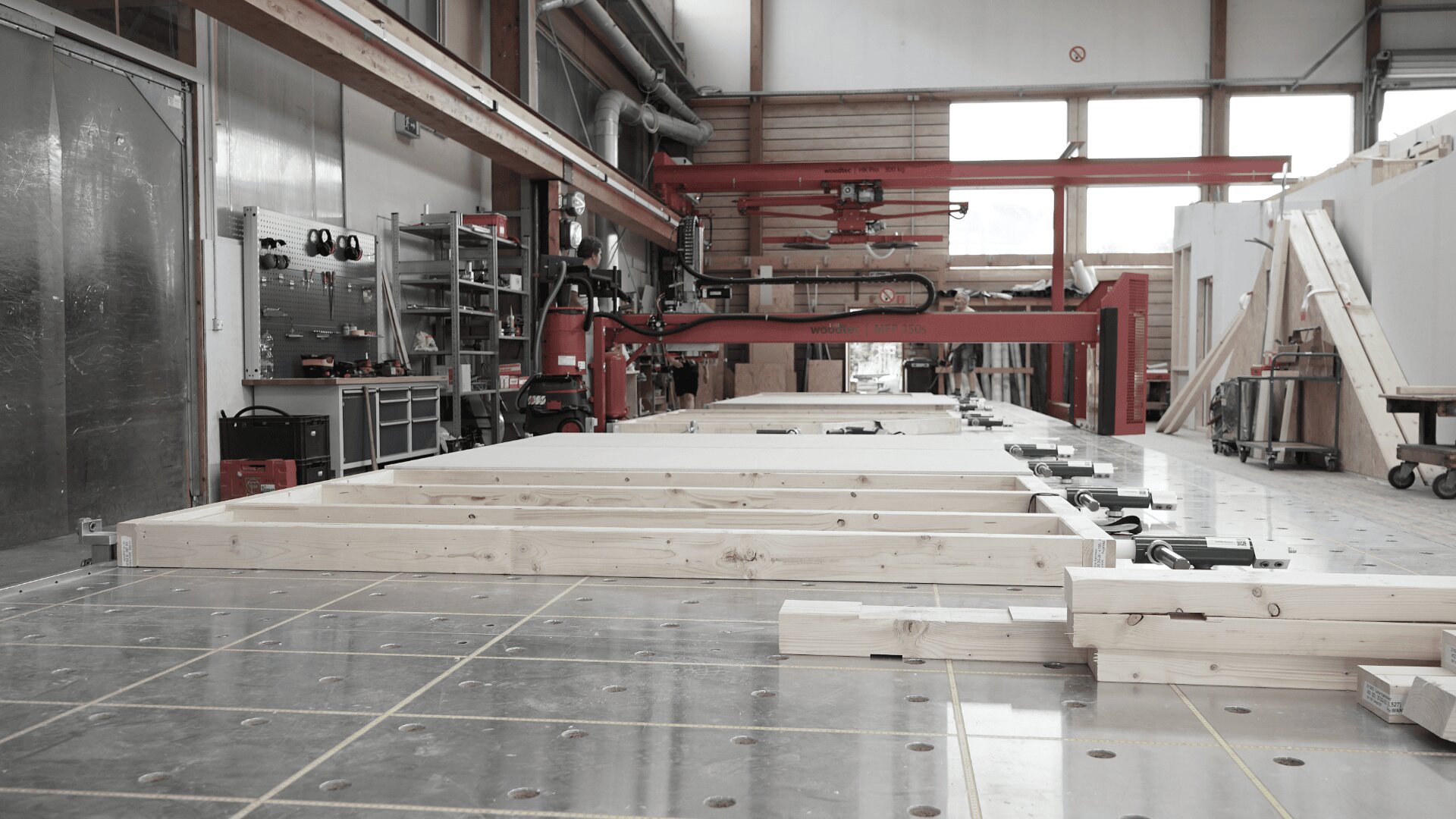 Eine moderne Holzverarbeitungsfabrik mit auf einem metallischen Arbeitstisch ausgelegten Holzbalken, umgeben von Industriemaschinen, Regalen mit Werkzeugen und großen Fenstern, die natürliches Licht hereinlassen.