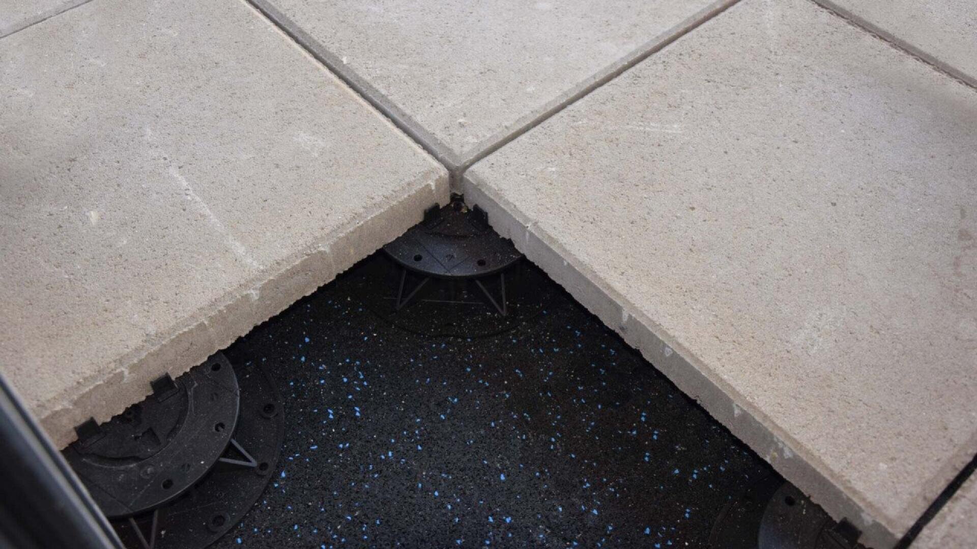 Four large concrete paving slabs are installed on adjustable plastic pedestals, with one slab removed to reveal the black support system and surface underneath, speckled with small blue dots.
