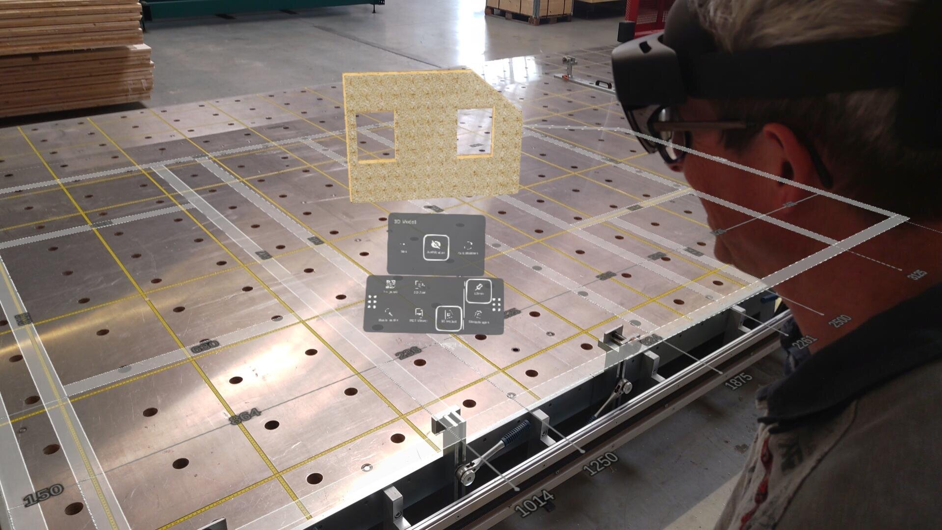 Eine Person mit Augmented-Reality-Brille sieht eine virtuelle Schnittstelle und ein 3D-Modell eines Hauses, das auf einen großen Metallgittertisch in einer industriellen Umgebung projiziert wird.