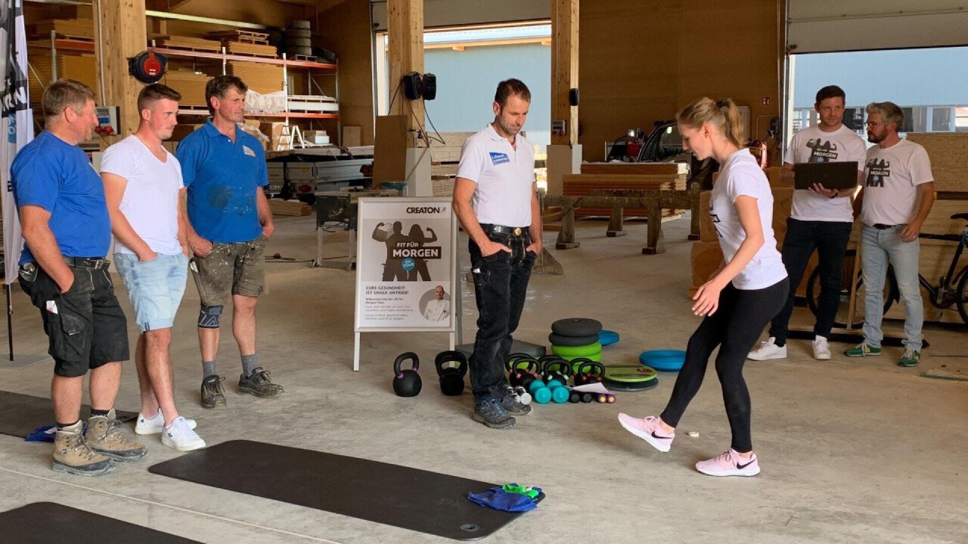 A woman in athletic clothing demonstrates an exercise on a mat to a group of men wearing work clothes in a spacious indoor workshop. Exercise equipment and wooden structures are visible in the background.