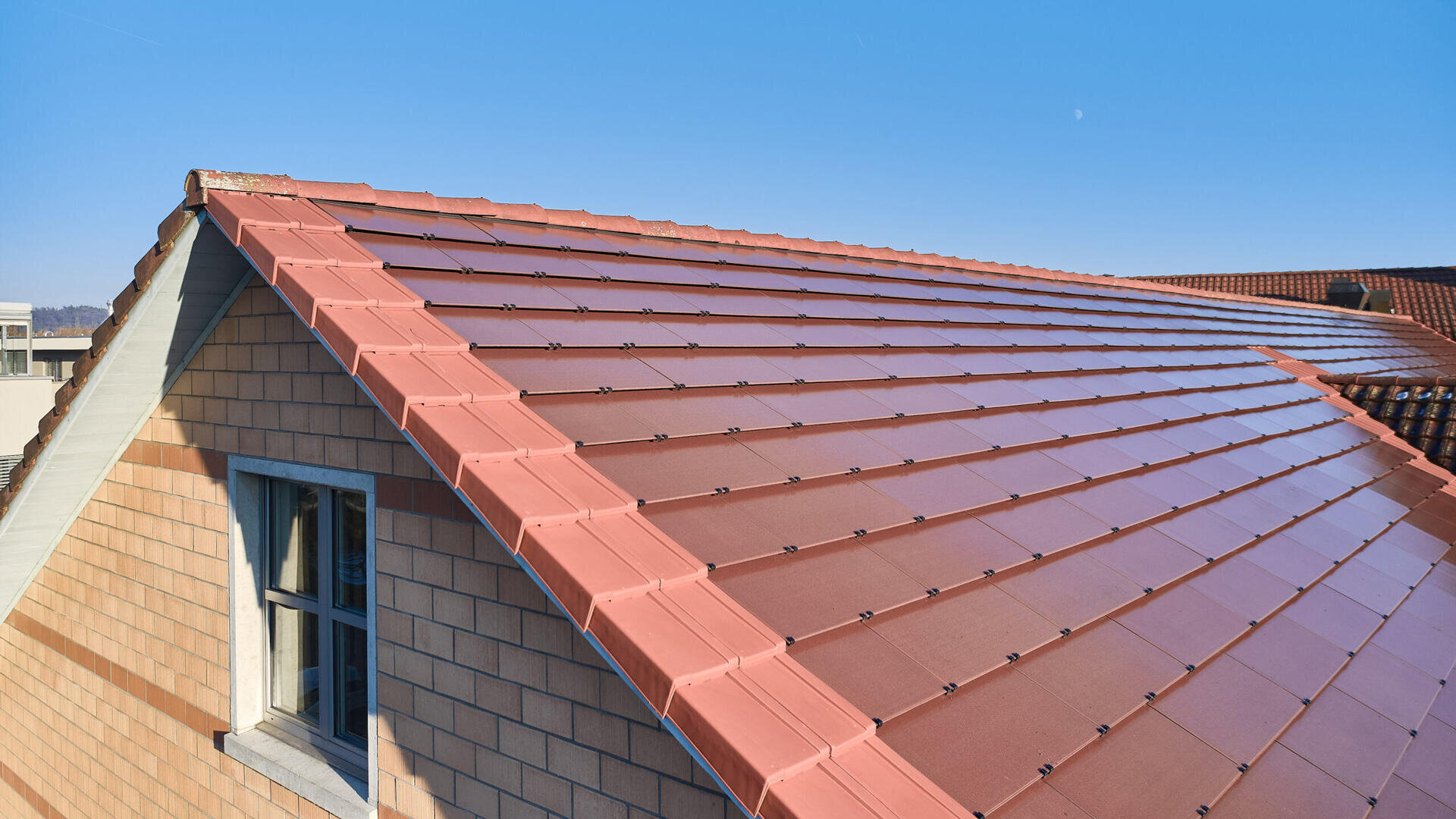 Unter einem klaren blauen Himmel ist ein mit roten Solarziegeln gedecktes Schrägdach zu sehen. Die Außenwand ist mit beigen und braunen Ziegeln verkleidet, und auf der linken Seite ist ein kleines Fenster zu sehen.