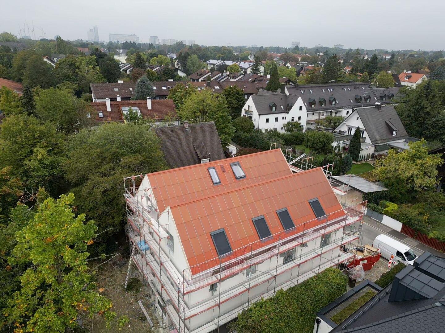 Aerial view of a house under construction with scaffolding and a partially completed red roof, surrounded by trees and neighboring houses in a suburb.