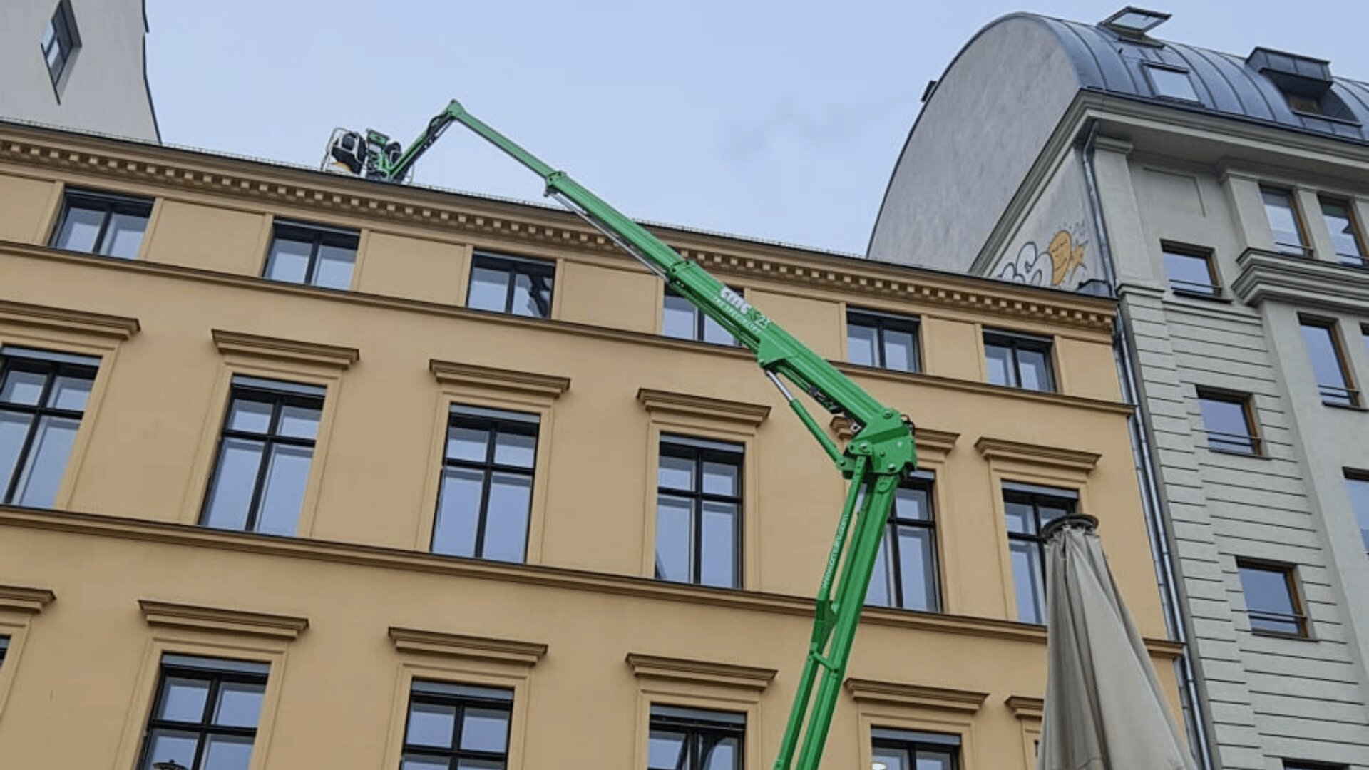 Eine grüne Hebebühne fährt auf das Dach eines beigefarbenen Gebäudes, wo ein Arbeiter in einem Sicherheitskäfig die Kante zu inspizieren oder zu reparieren scheint. Angrenzende Gebäude und Fenster sind unter einem bewölkten Himmel sichtbar.