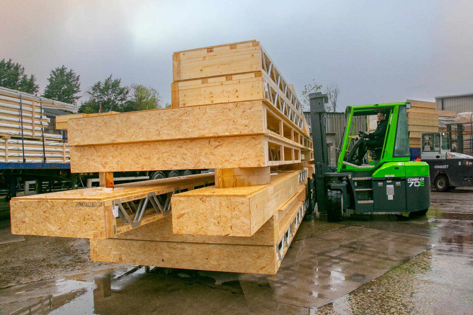 Ein grüner Gabelstapler hebt an einem bewölkten Tag einen großen Stapel von Holzbalken und -platten auf einem Industriegelände an. Der nasse Boden deutet auf kürzlichen Regen hin; andere Holzstapel sind im Hintergrund zu sehen.