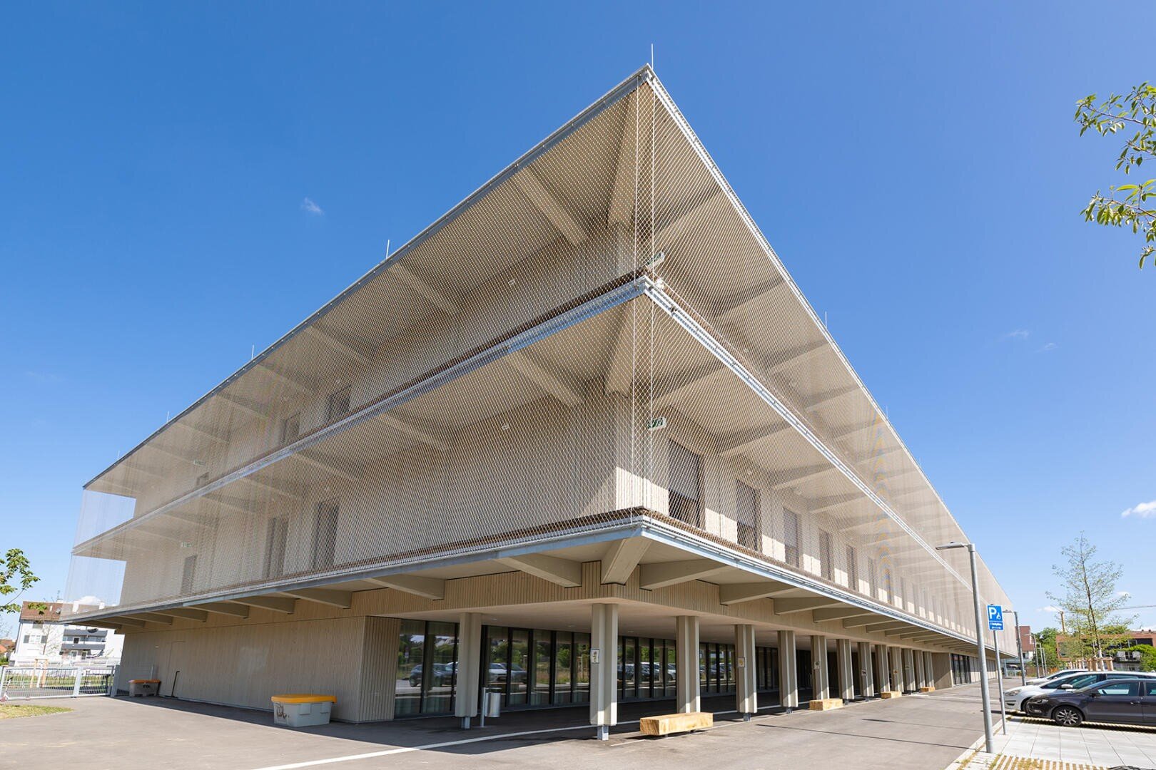 Ein modernes, dreistöckiges Gebäude mit offenen Balkonen und einer netzartigen Außenfassade vor einem strahlend blauen Himmel. Das Erdgeschoss verfügt über Glaseingänge und Parkplätze in der Nähe.