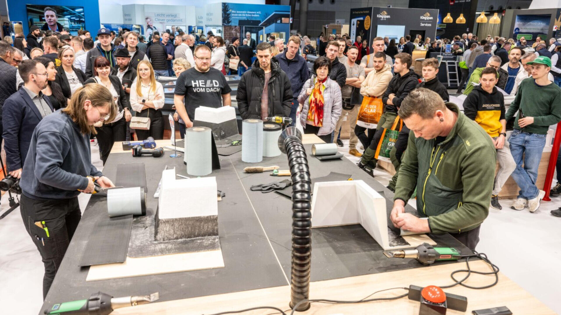 Eine Menschenmenge beobachtet zwei Personen, die auf einem Messe- oder Ausstellungsstand Bautechniken mit Dämmblöcken und Werkzeug vorführen. Verschiedene Zuschauer um den Tisch herum beobachten aufmerksam.
