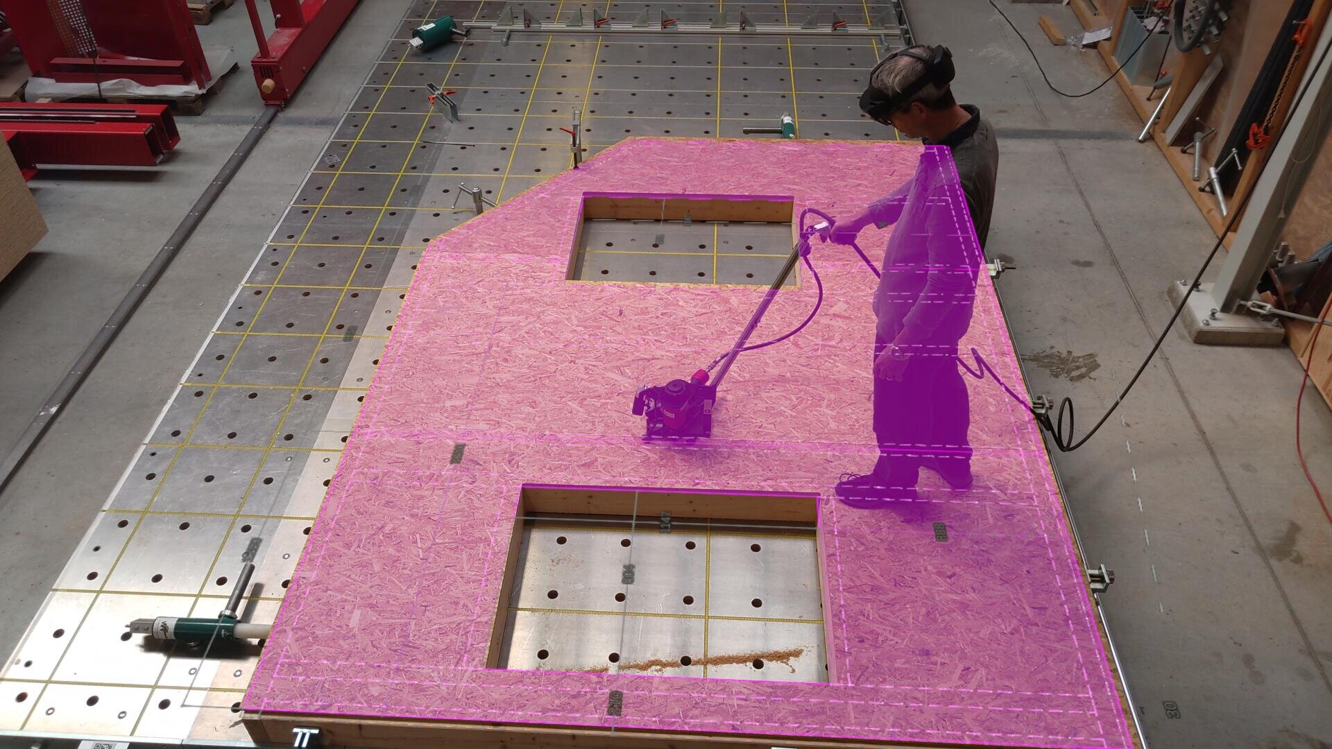 Eine Person interagiert mithilfe eines VR-Headsets und eines Controllers mit einer großen Holzplatte auf dem Boden einer Werkstatt. Teile der Platte und die Person selbst sind mit durchscheinenden violetten digitalen Grafiken überlagert.