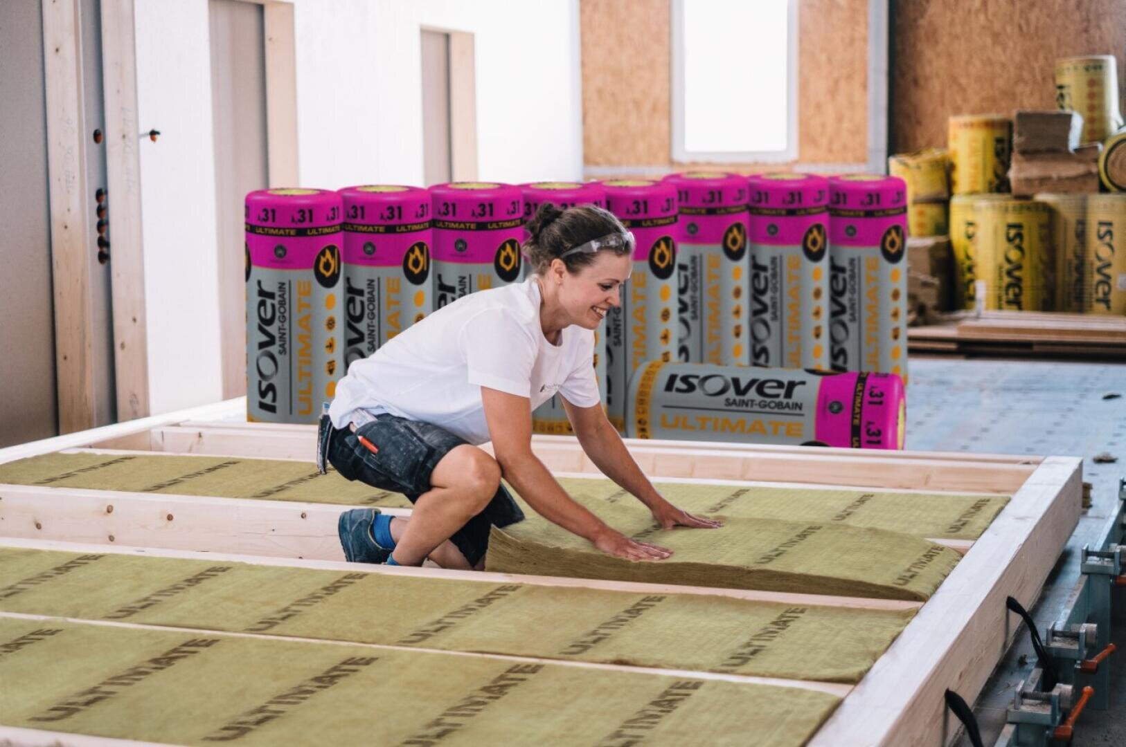 Eine Person in Arbeitskleidung bringt in einem im Bau befindlichen Gebäude Dämmplatten zwischen Holzbalken an. Im Hintergrund sind Stapel von Dämmmaterial zu sehen.