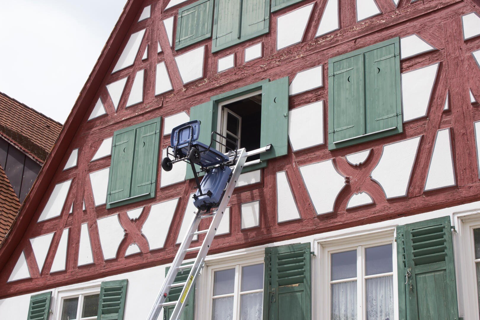 An elevator pulls up to an open window on the second floor of a traditional half-timbered house with green shutters and decorative red wooden beams.