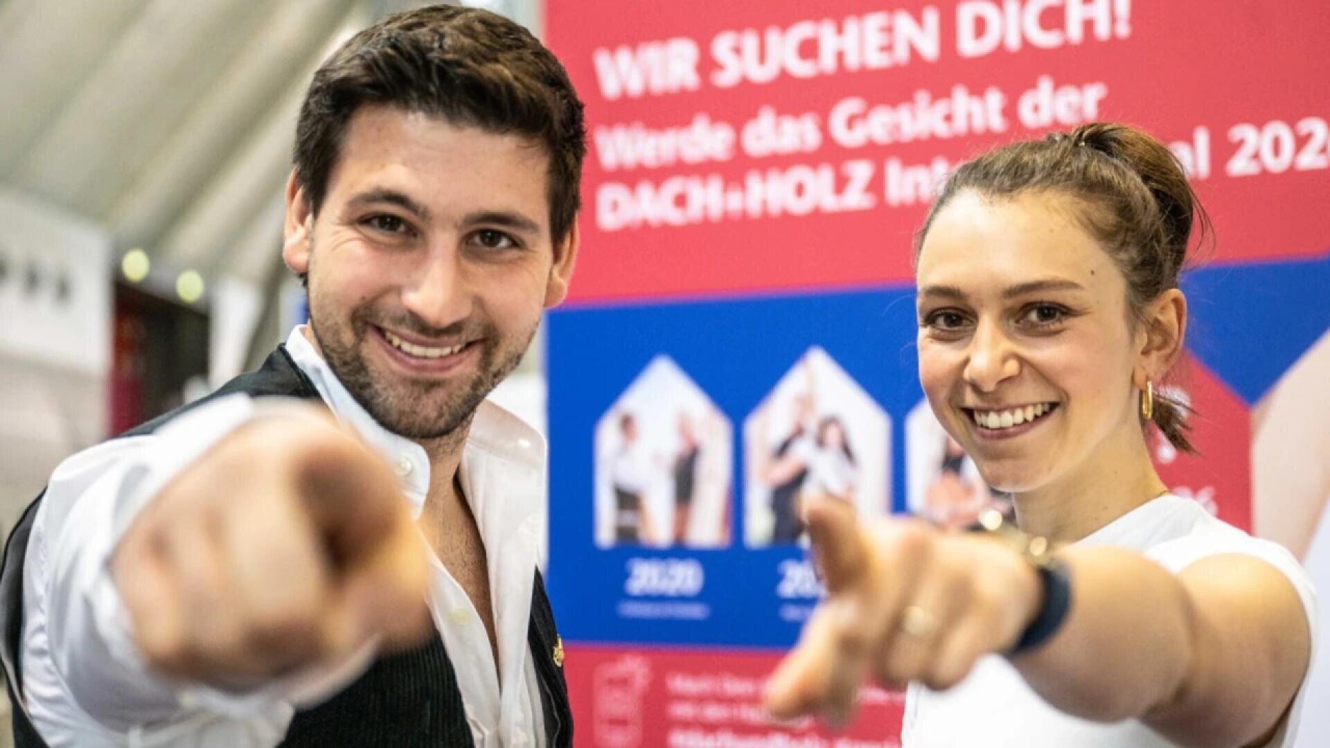 A smiling man and woman stand in front of a red and blue sign, pointing toward the camera. The background has German text, including Wir suchen dich! in white letters.