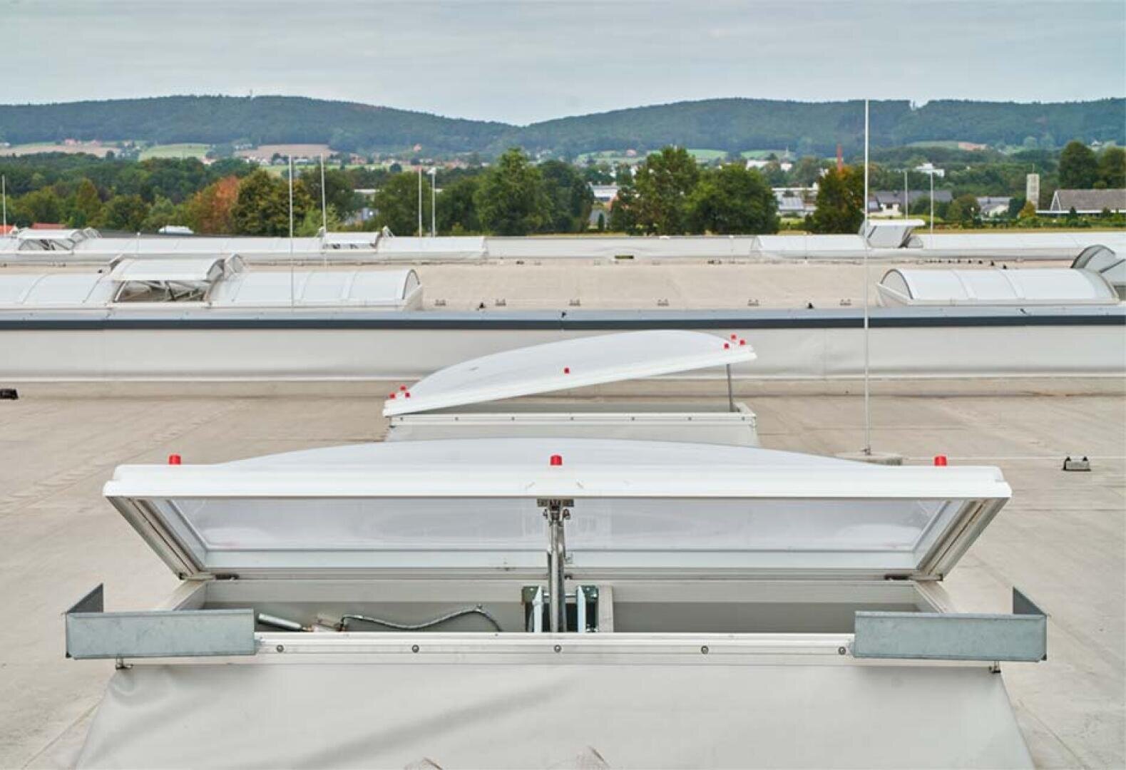 Ein flaches Industriedach mit mehreren rechteckigen weißen Schornsteinen, einige offen, andere geschlossen, umgeben von Bäumen und fernen Hügeln unter einem bewölkten Himmel.