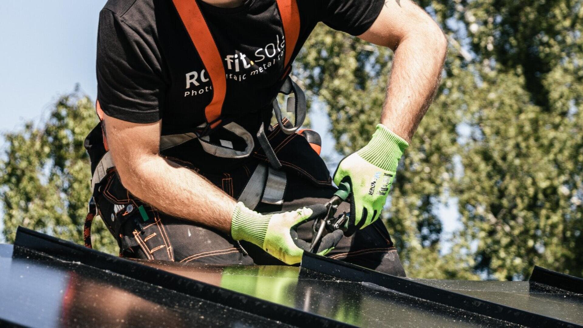 A person wearing safety gear and gloves installs solar panels on a roof, using a tool to secure the panels. Trees are visible in the background.