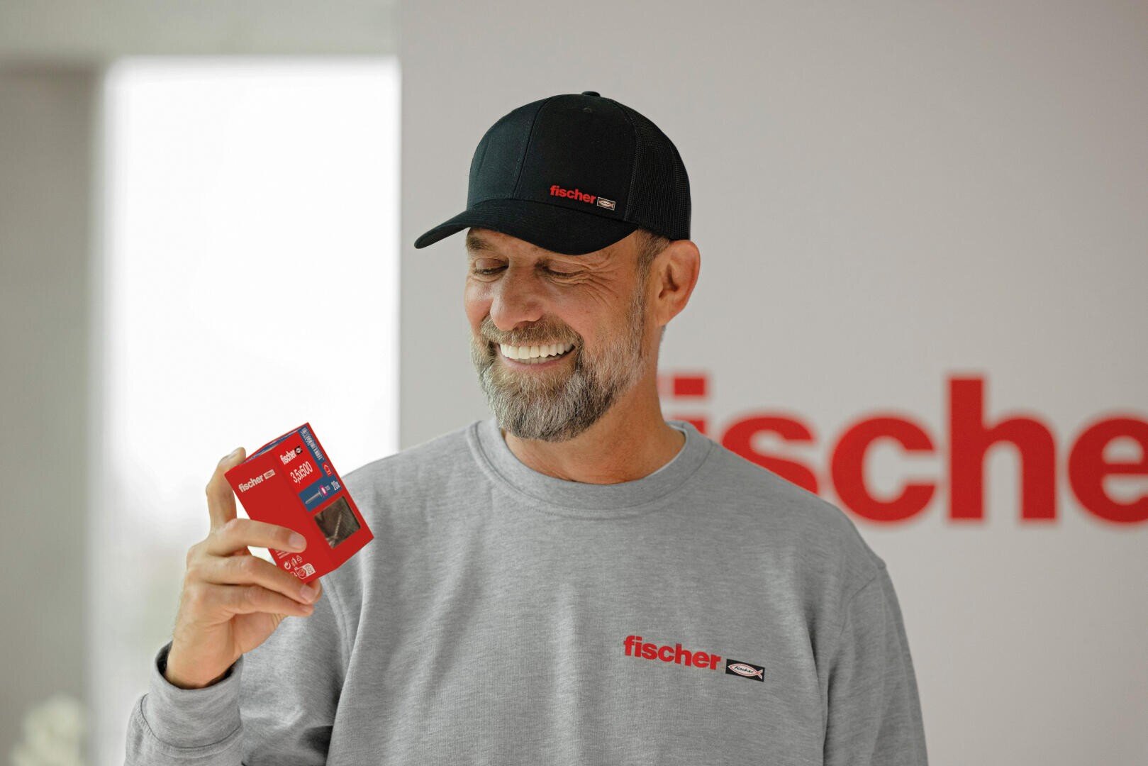 A smiling man in a fisherman's cap and sweatshirt holds a small red box. A blurred wall can be seen in the background, on which the word fischer can be partially read in red letters.