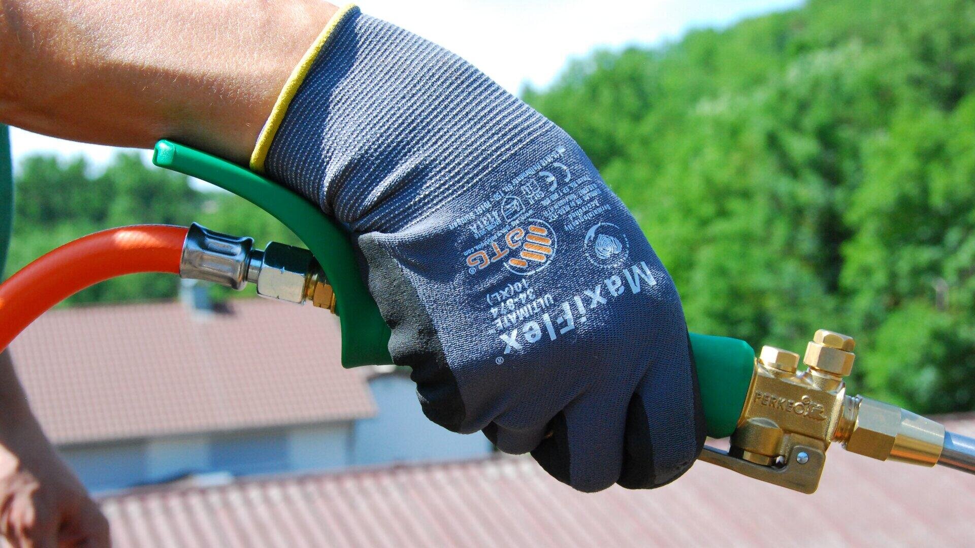 Eine behandschuhte Hand hält eine grüne und messingfarbene Lötlampe mit einem orangefarbenen Schlauch, im Freien, mit einem unscharfen Hintergrund aus Dächern und grünen Bäumen.