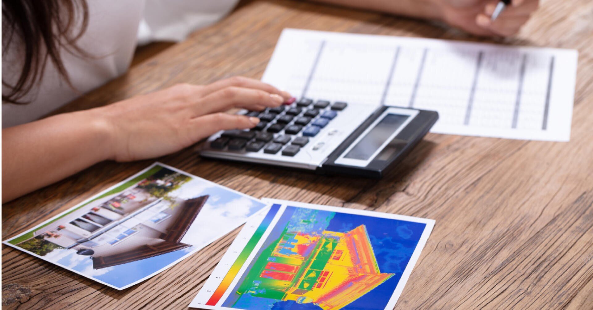 A person uses a calculator next to a document and two photographs, one of a house and one showing a thermal image of a building, on a wooden table.