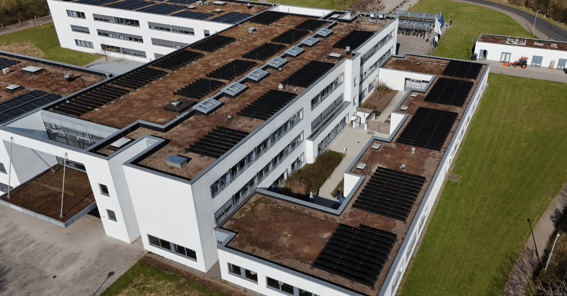 Luftaufnahme eines modernen weißen Gebäudes mit mehreren Stockwerken, einem mit Sonnenkollektoren und Oberlichtern bedeckten Flachdach, umgeben von Rasen und gepflasterten Wegen.