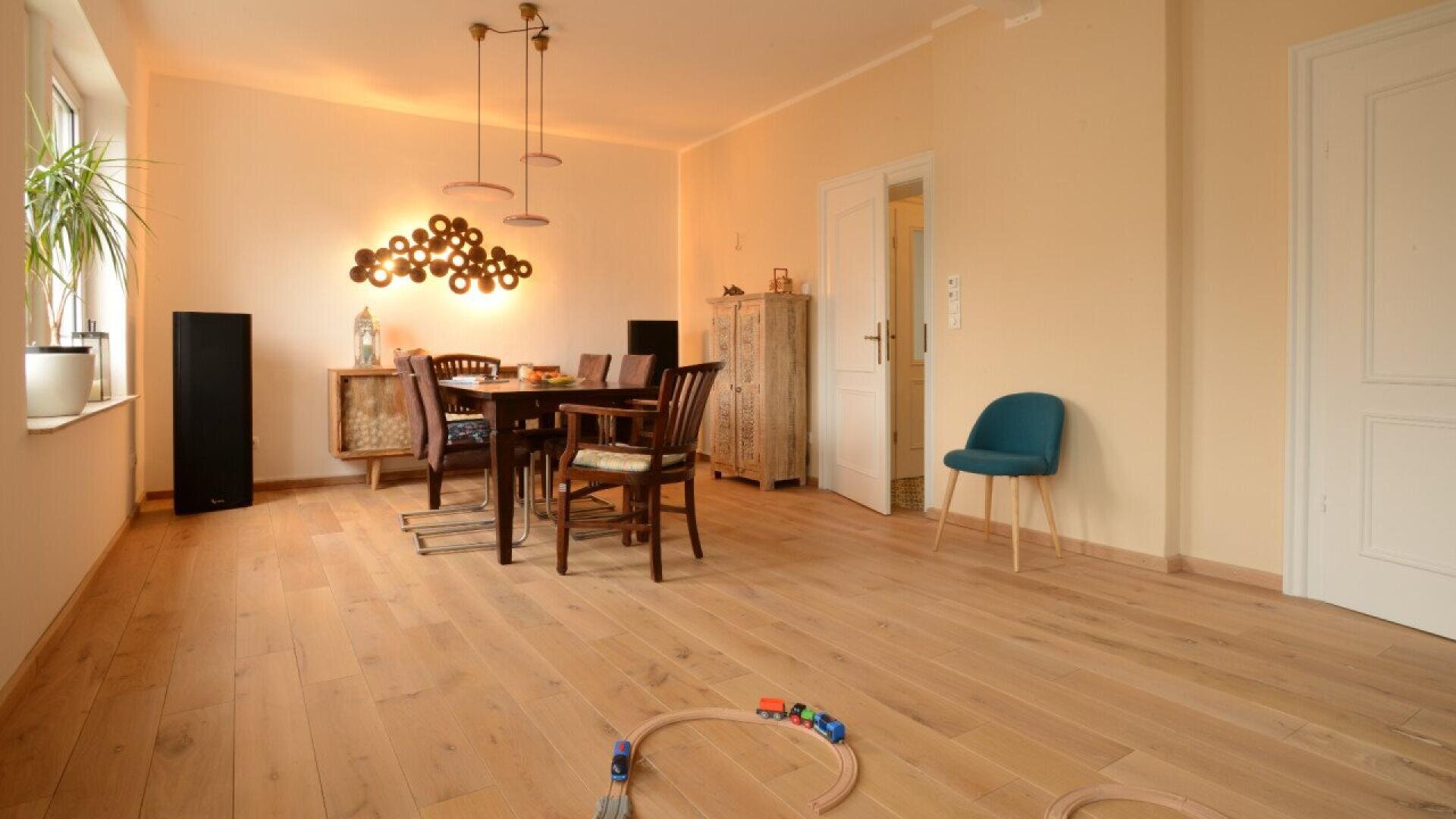 A dining room with wooden flooring, a dining table and chairs, a blue chair, a decorative cabinet, modern light fixtures, houseplants, and a toy train set on the floor in the foreground.