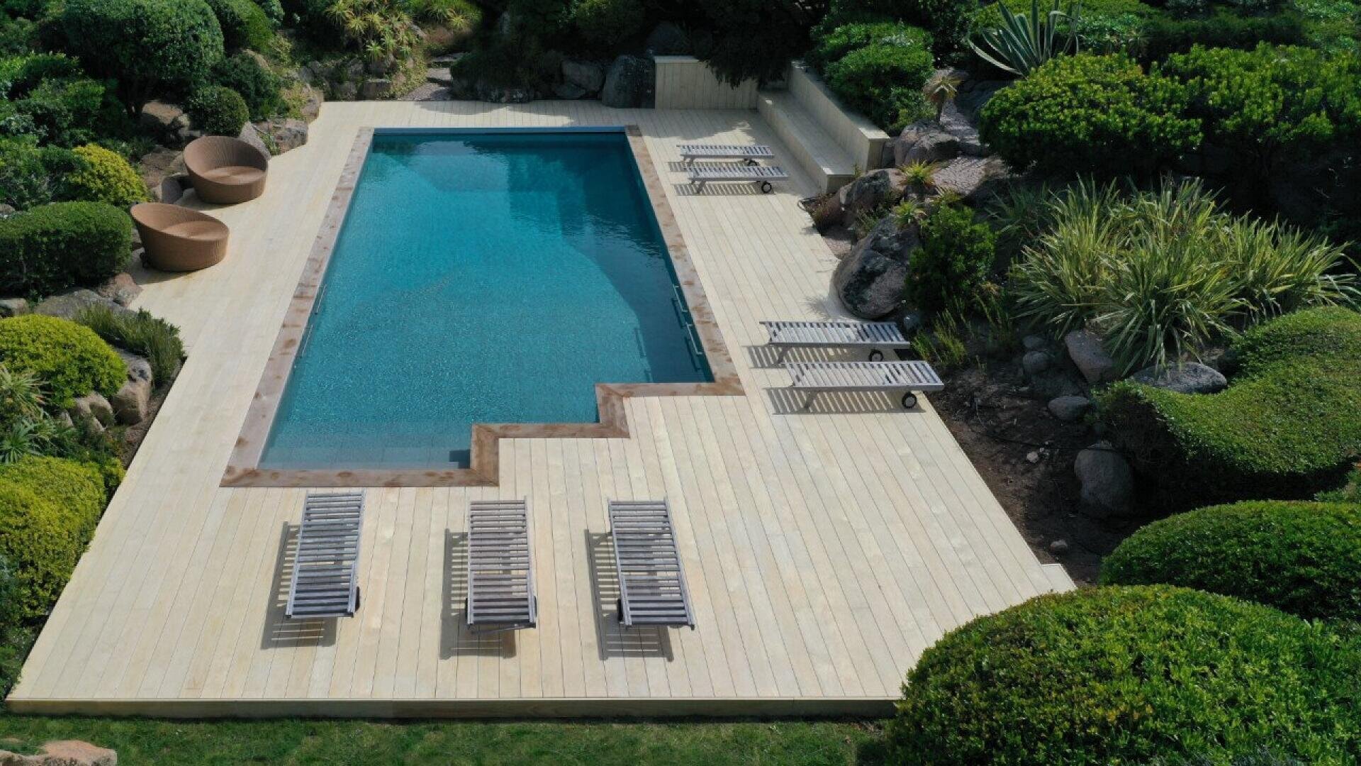 A rectangular outdoor swimming pool with a light wooden deck, surrounded by green bushes. Several lounge chairs and two round chairs are placed on the deck near the pool.