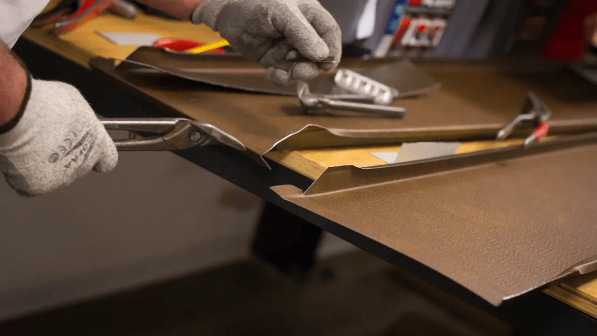 Eine Person mit Handschuhen schneidet mit einer Blechschere ein braunes Metallblech auf einer Werkbank zu. In der Nähe liegen verstreut verschiedene Werkzeuge wie ein Hammer und eine Zange.