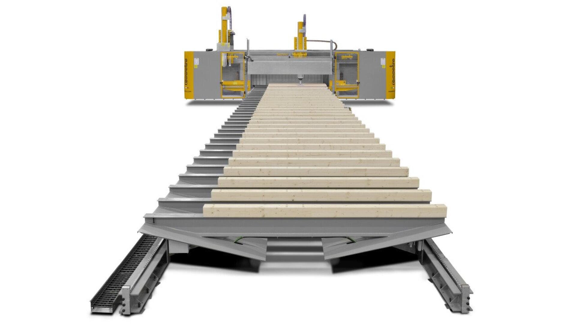 Front view of an automated industrial machine laying multiple wooden beams in parallel rows on a metal frame, with yellow mechanical arms visible in the background.