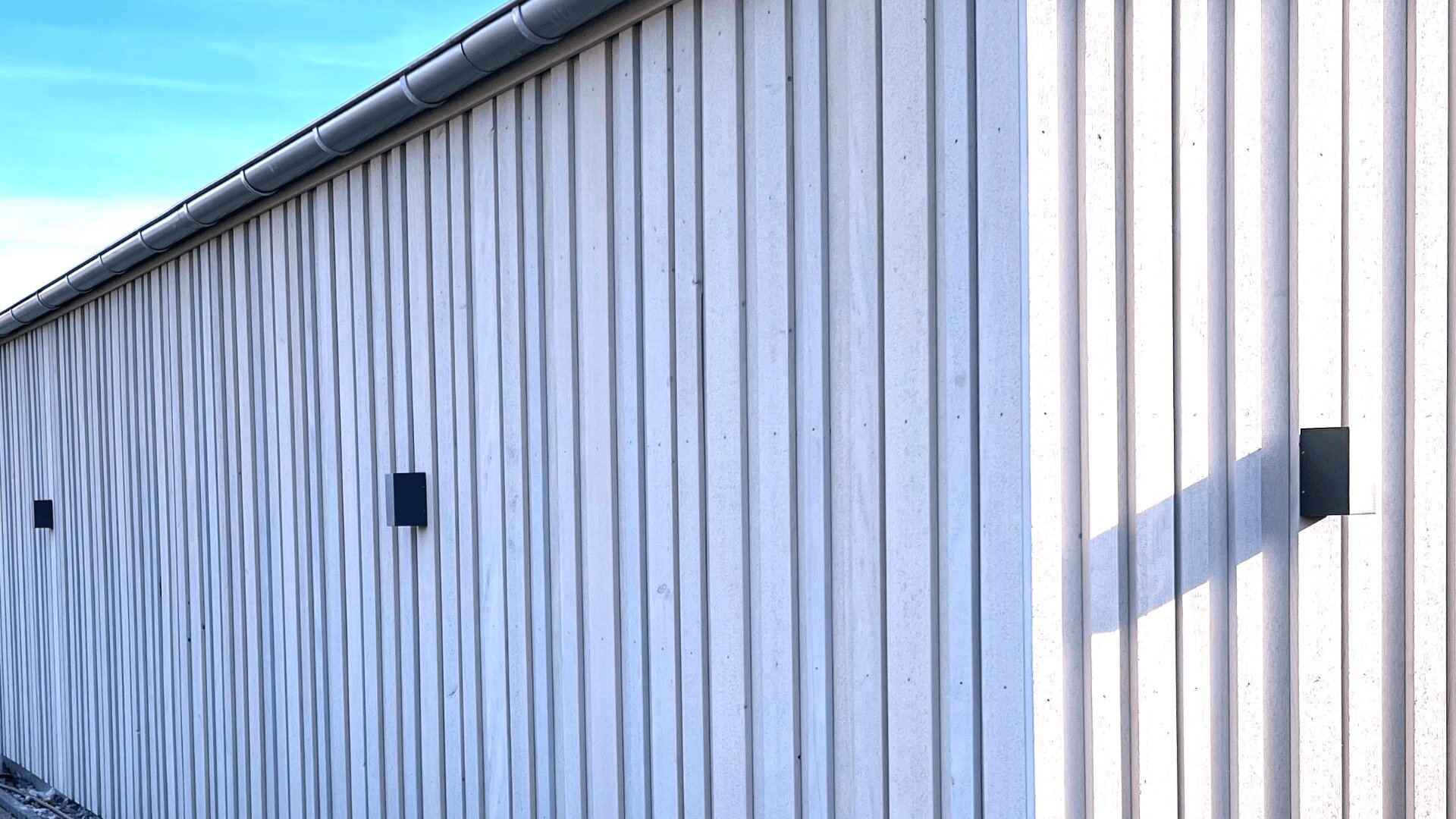 Eine hellgraue Holzwand mit vertikalen Paneelen, drei schwarze rechteckige Wandleuchten, die Schatten werfen, und eine Dachrinne, die unter einem blauen Himmel entlang des Daches verläuft.