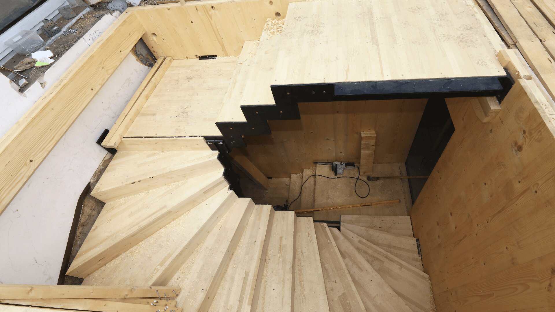 Eine teilweise fertiggestellte Holztreppe mit unfertigen Stufen, schwarzen Metallstützen und Bauwerkzeugen auf einer unteren Ebene in einem Gebäude, das gerade renoviert wird.