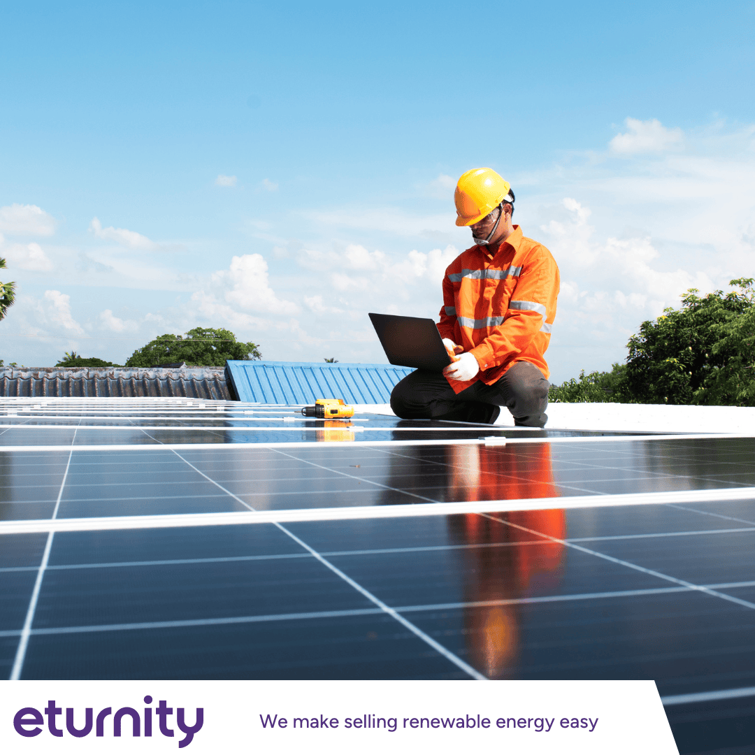 Ein Arbeiter in orangefarbener Sicherheitsweste und Helm kniet auf einem Solarpanel auf dem Dach und nutzt seinen Laptop unter blauem Himmel. Der Text lautet: Ewigkeit – Wir machen den Verkauf erneuerbarer Energien einfach.