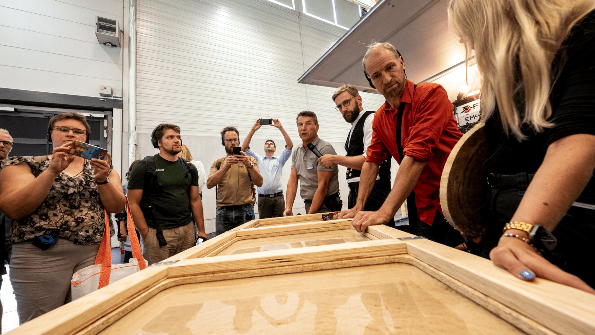 A group of people, some taking photos, gather around a man in a red shirt who is explaining a wooden frame structure on a table in a bright indoor setting.
