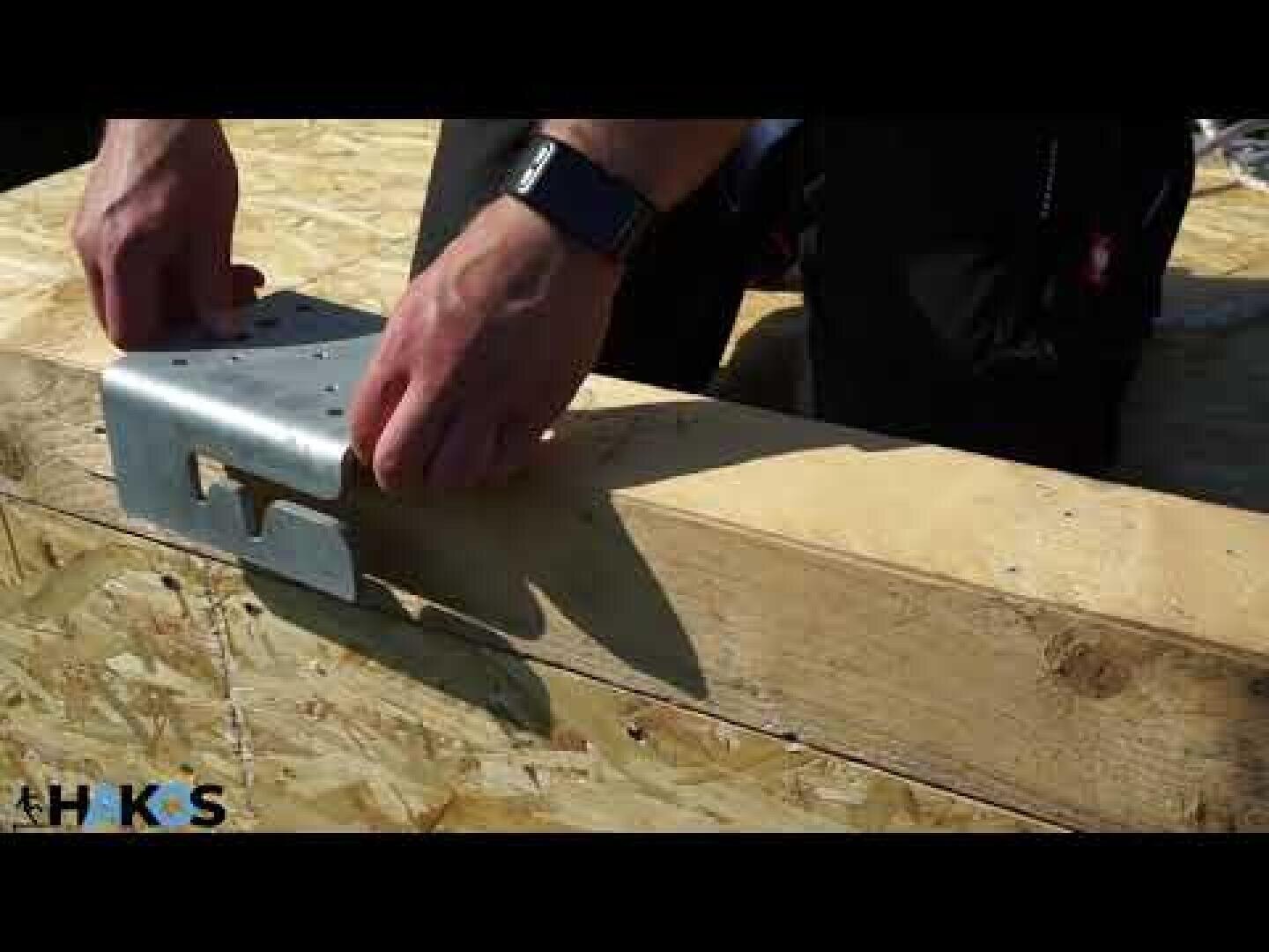A person wearing a smartwatch attaches a metal connector to a wooden beam and prepares to build. The wood has visible screws and the scene is illuminated by natural daylight.