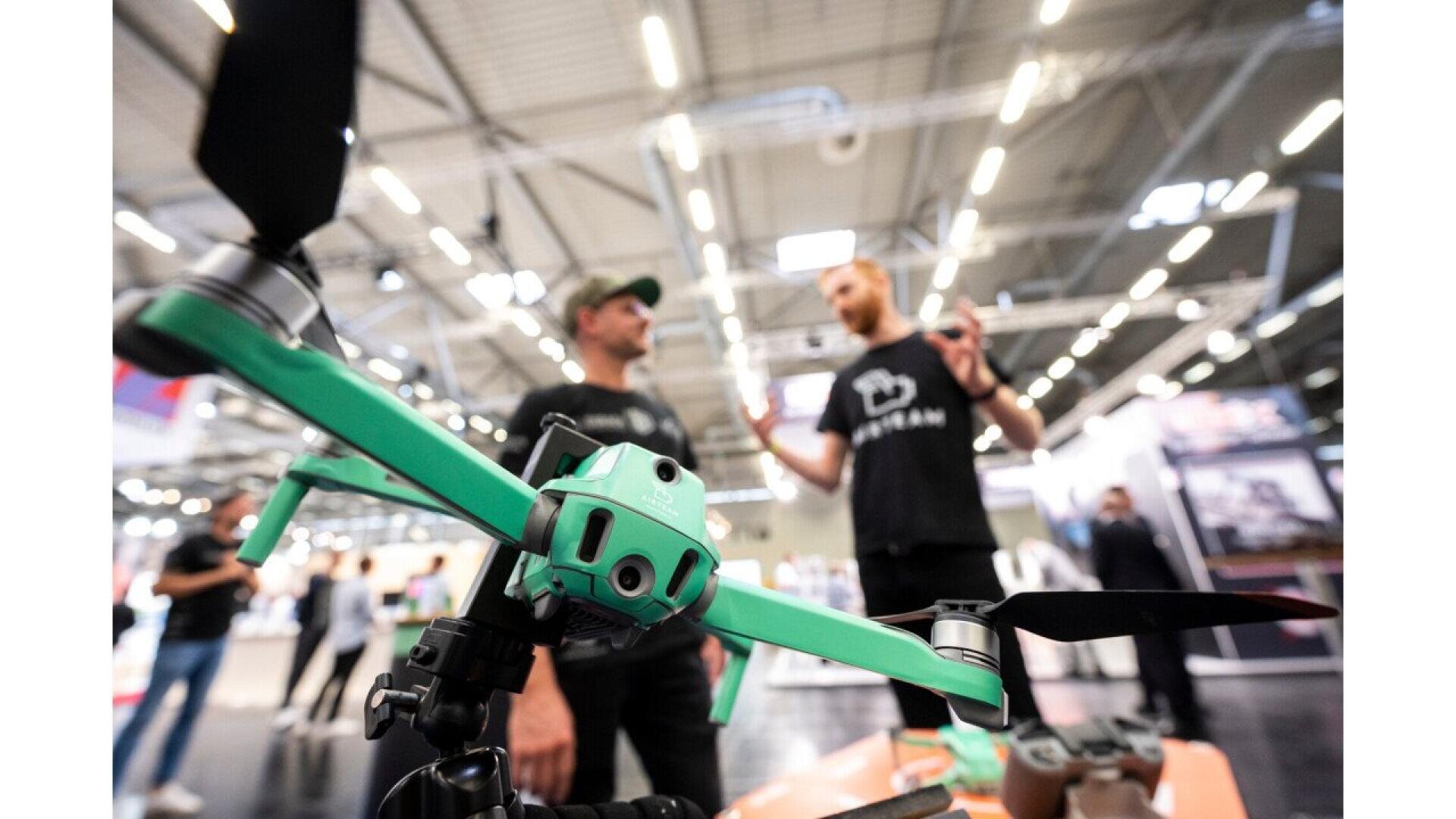 A bright green drone is displayed prominently in the foreground, while two men in black shirts have a conversation in the blurred background at an indoor event or expo.
