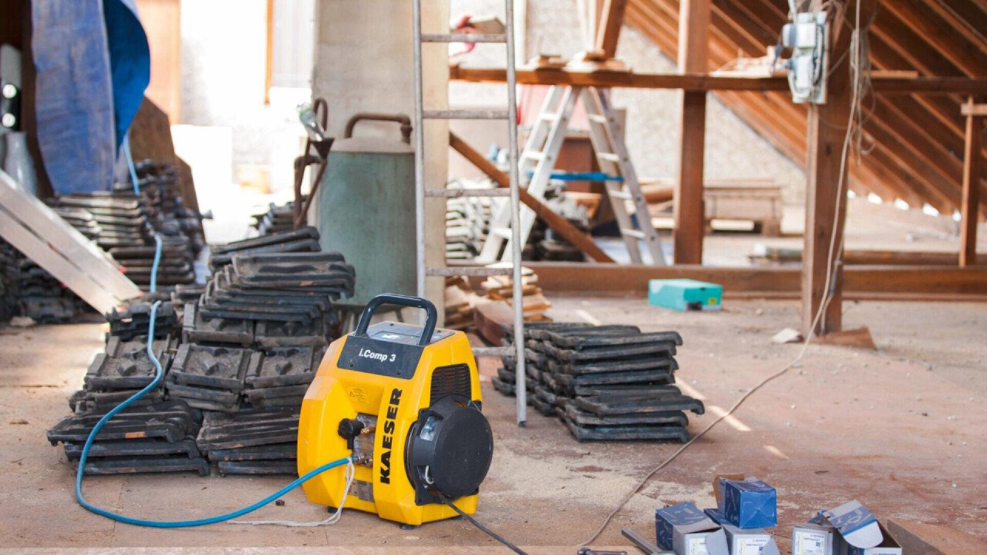 A yellow KAESER air compressor sits on a dusty floor in a construction area, surrounded by stacked black tiles, cables, and tools, with ladders and exposed beams visible in the background.