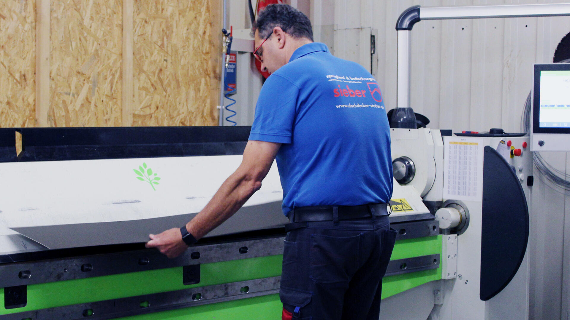 Ein Mann im blauen Hemd bedient eine große industrielle Biegemaschine im Innenbereich und hält ein Blech mit einem grünen Blattlogo in der Hand. Auf der rechten Seite sind ein Monitor und ein Bedienfeld zu sehen.