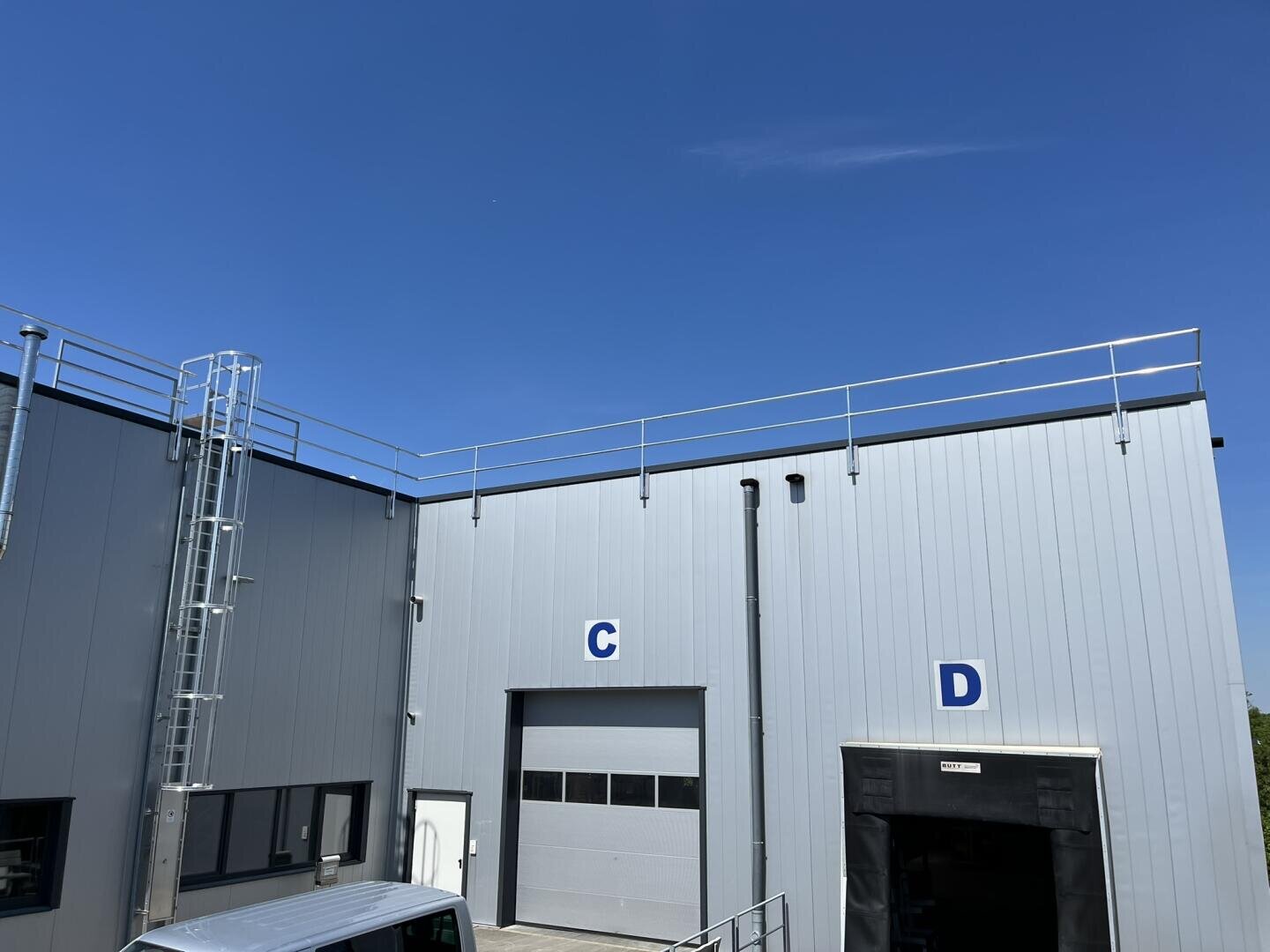 Ein modernes Industriegebäude mit grauen Metallverkleidungen, beschrifteten Abschnitten C und D, Garagentoren, einer vertikalen Leiter und Sicherheitsgeländern auf dem Dach vor einem klaren blauen Himmel.