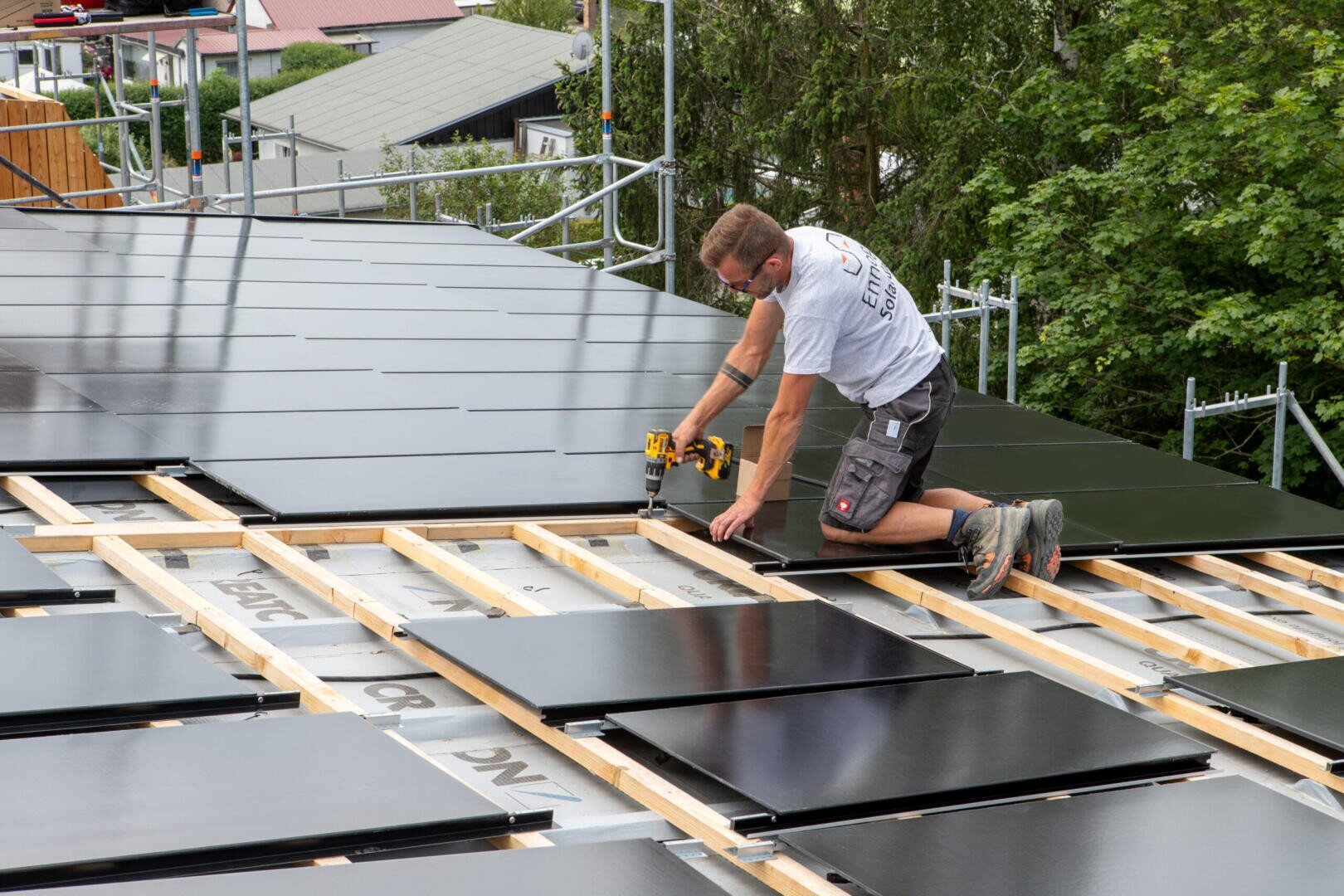 A person kneels on a sloping roof and installs solar cells with a drill. The wooden scaffolding supports the panels, and trees and buildings can be seen in the background.