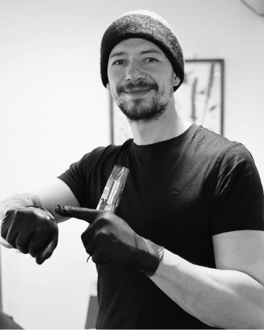 Ein Mann mit schwarzer Mütze, schwarzem T-Shirt und schwarzen Handschuhen lächelt, während er eine Tätowiermaschine in der Hand hält und in einem Raum mit gerahmten Kunstwerken im verschwommenen Hintergrund steht.