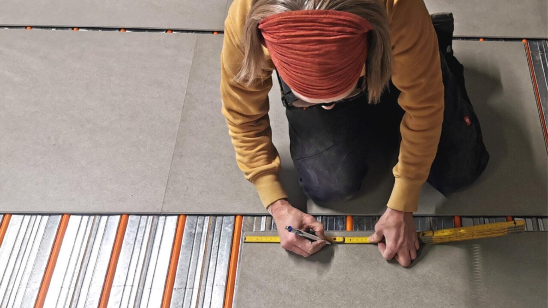 A person kneels on the floor, marking a large tile with a pencil and measuring tape. Metal heating strips are visible beneath the tile, indicating flooring installation work.