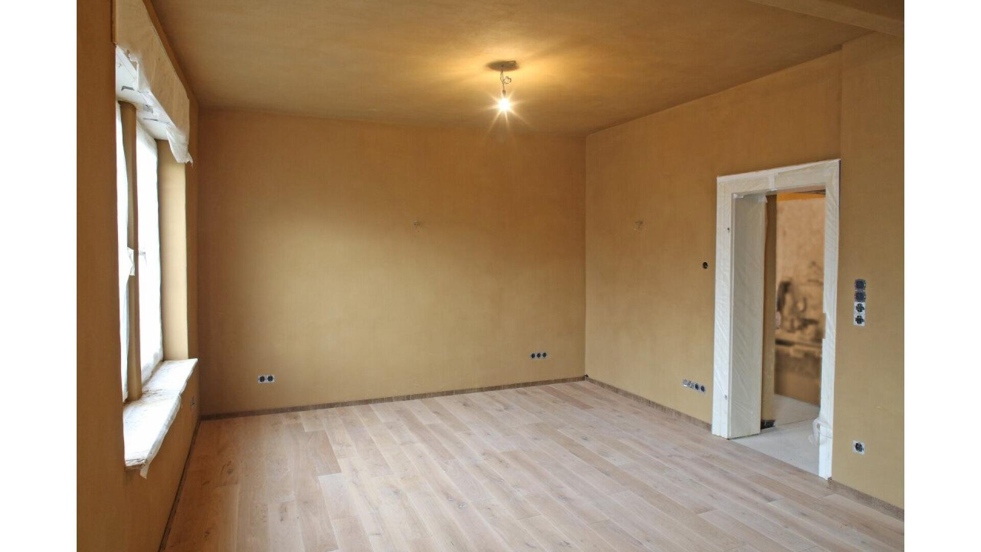 An empty room with light wooden flooring, tan-colored walls, a single ceiling light, two windows on the left, and an open doorway on the right leading to another room.