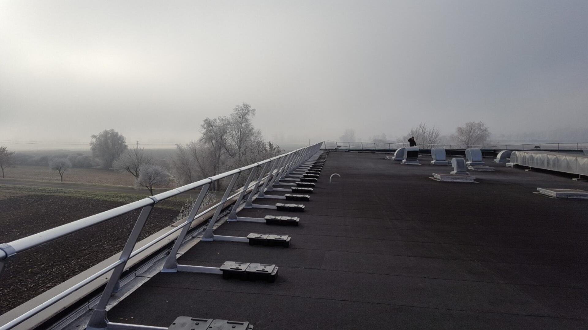 A flat roof with metal railings and ventilation systems, overlooking a misty landscape of bare trees and fields. A person stands near the edge in the distance, under an overcast sky.
