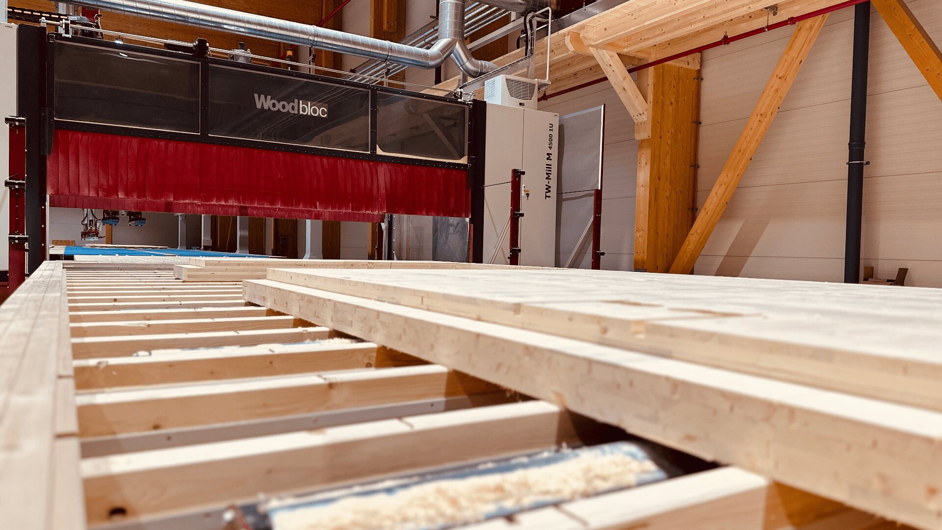 Eine große automatisierte Holzschneidemaschine mit der Bezeichnung Woodbloc verarbeitet Holzbalken in einer gut beleuchteten Industriewerkstatt mit freiliegenden Holzstützen und Lüftungsschächten.