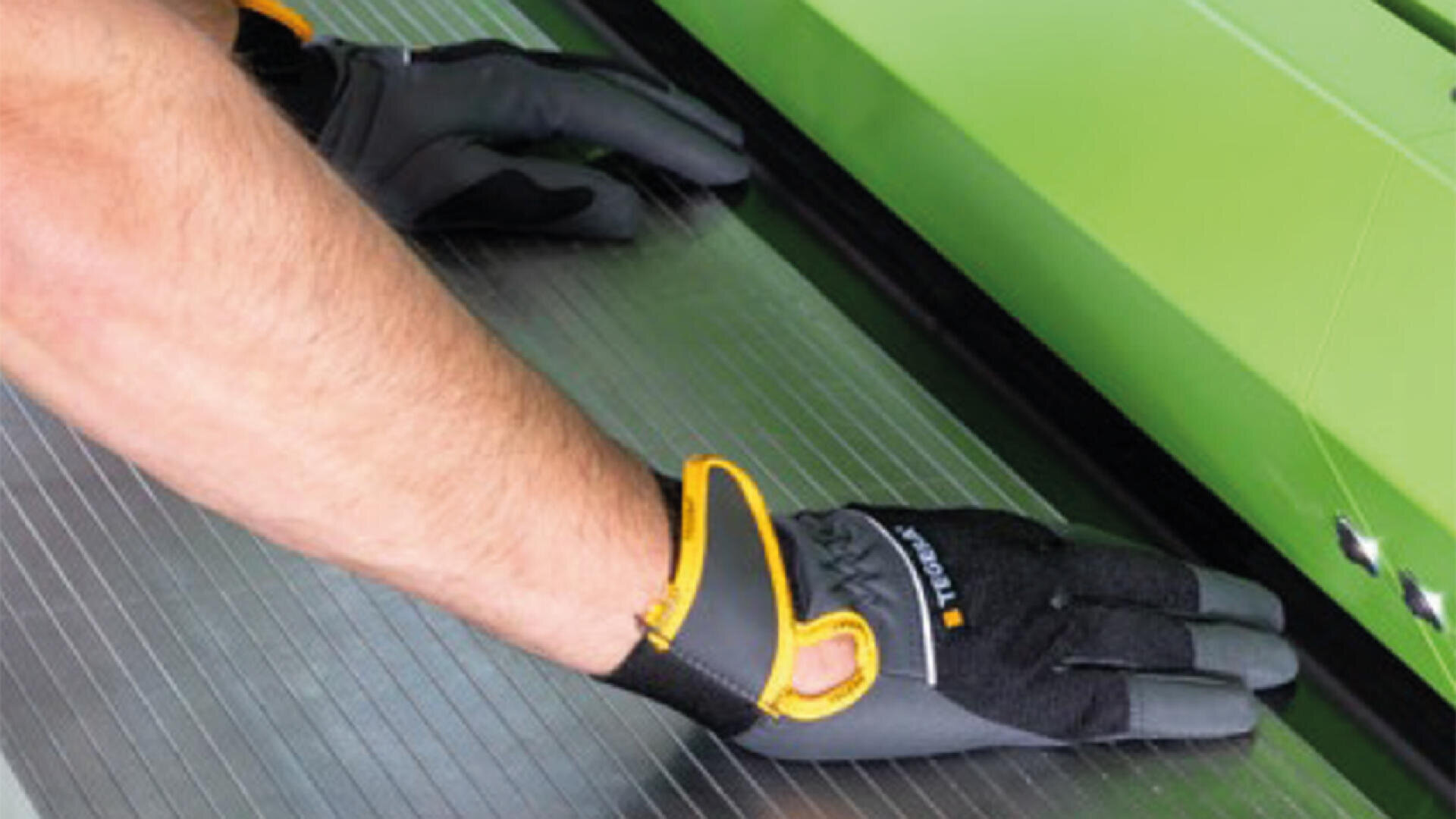 Nahaufnahme einer Person mit Schutzhandschuhen, die eine grüne Industriemaschine bedient und Material auf einer geriffelten Metalloberfläche ausrichtet oder führt.