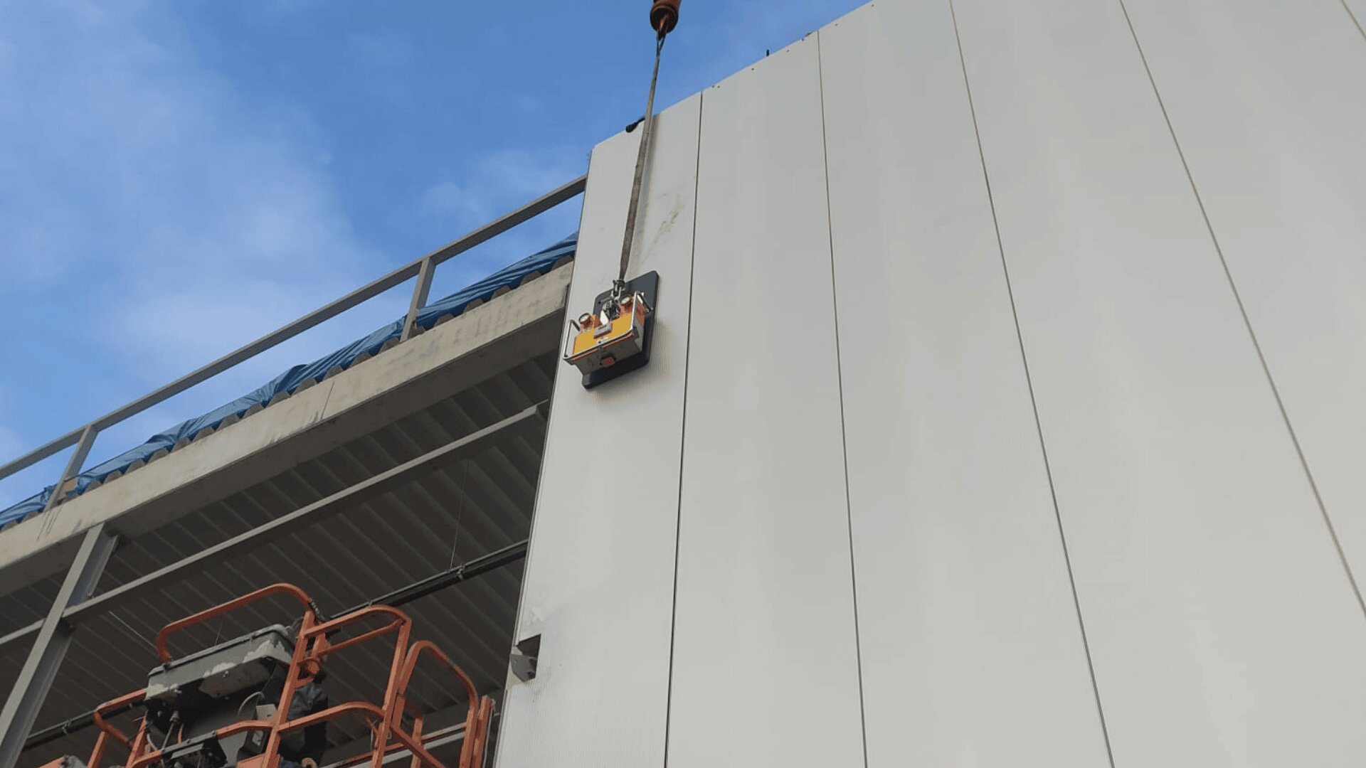 A large wall panel is being lifted by a crane next to an industrial building under a blue sky. Construction equipment is visible below.