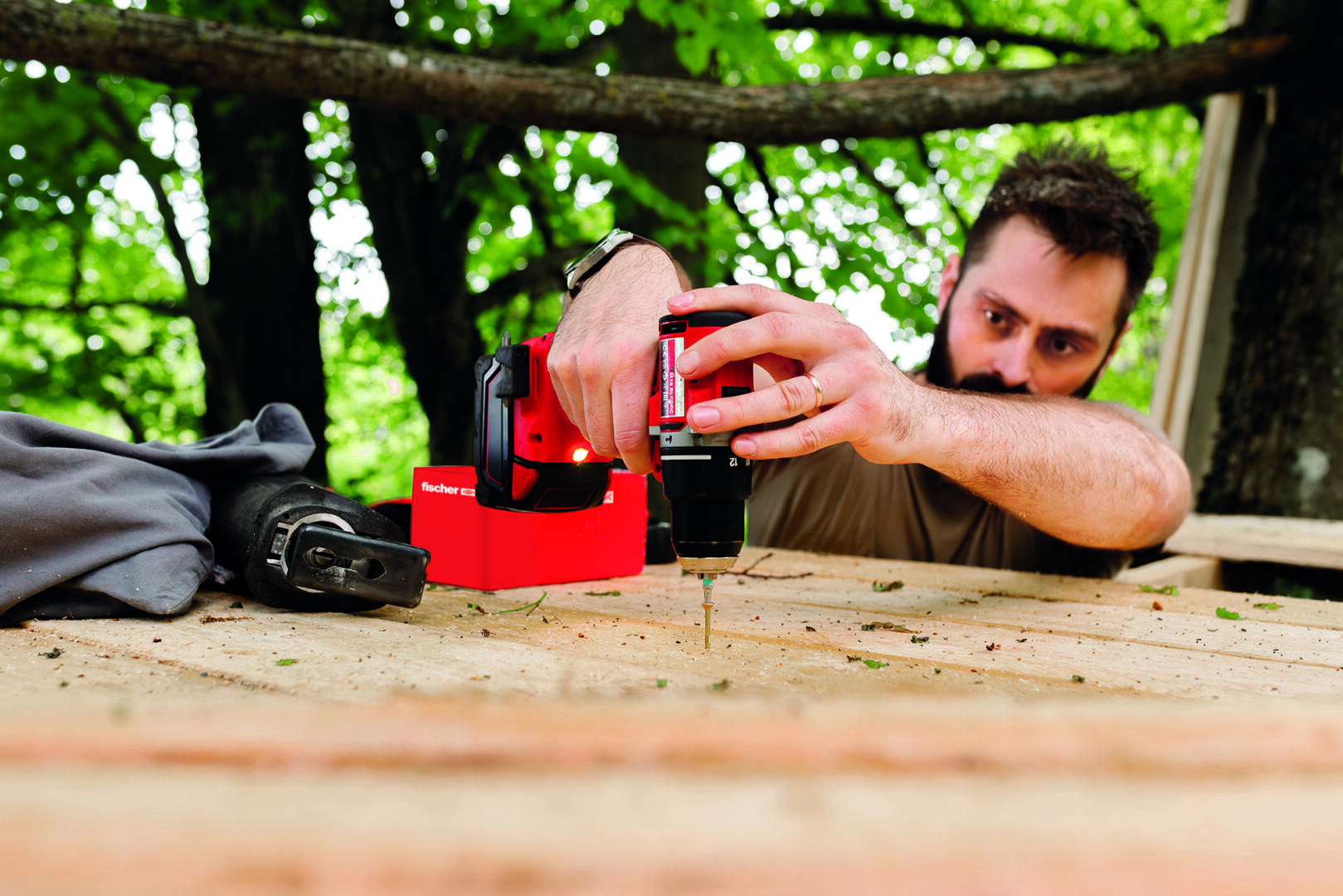 A man is using a drill to screw into a wooden surface outdoors, with trees and greenery visible in the background. He is concentrating on his task and the tools are placed nearby.