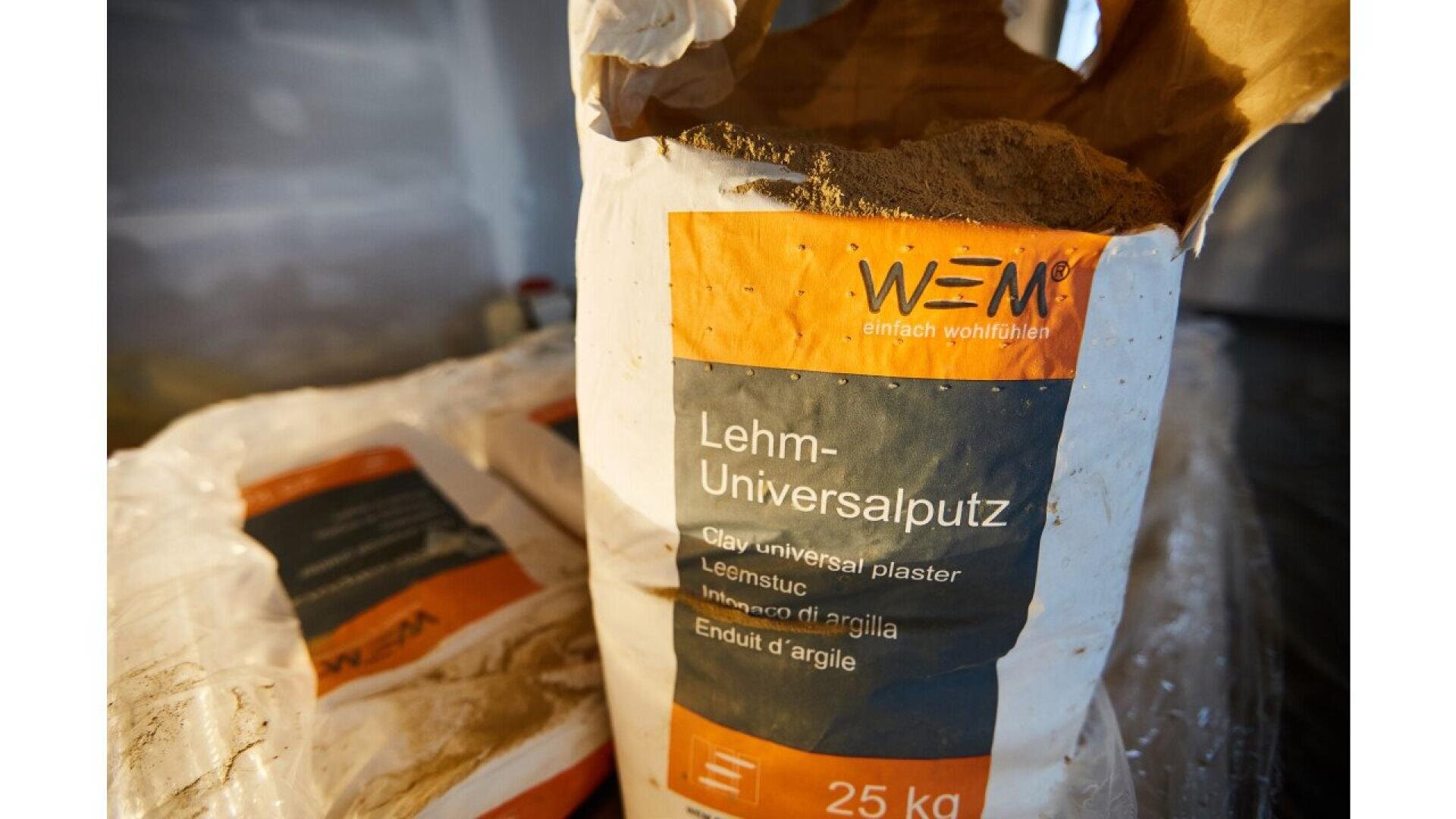 A close-up of an open 25 kg bag of WEM clay universal plaster, labeled in multiple languages, with another similar bag in the background.